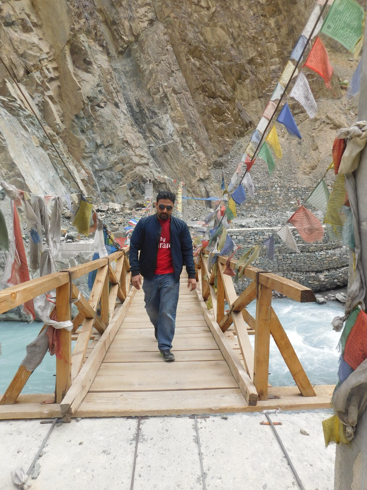 Vinaay Patil crossing a wooden suspension bridge strung with prayer flags over a glacial river in Ladakh.